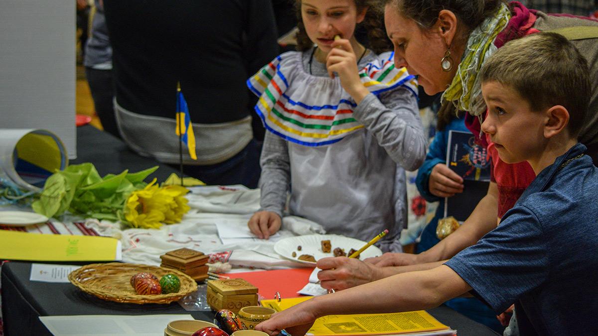 一小群人和孩子们检查装饰好的复活节彩蛋(Pysanky), faux sunflowers, 乌克兰套娃, 还有几个雕花木盒.  