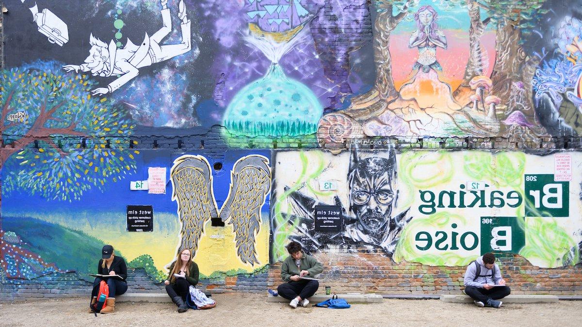 Four students sit on the ground sketching in front of a wall painted with colorful murals. One reads "Breaking Boise."